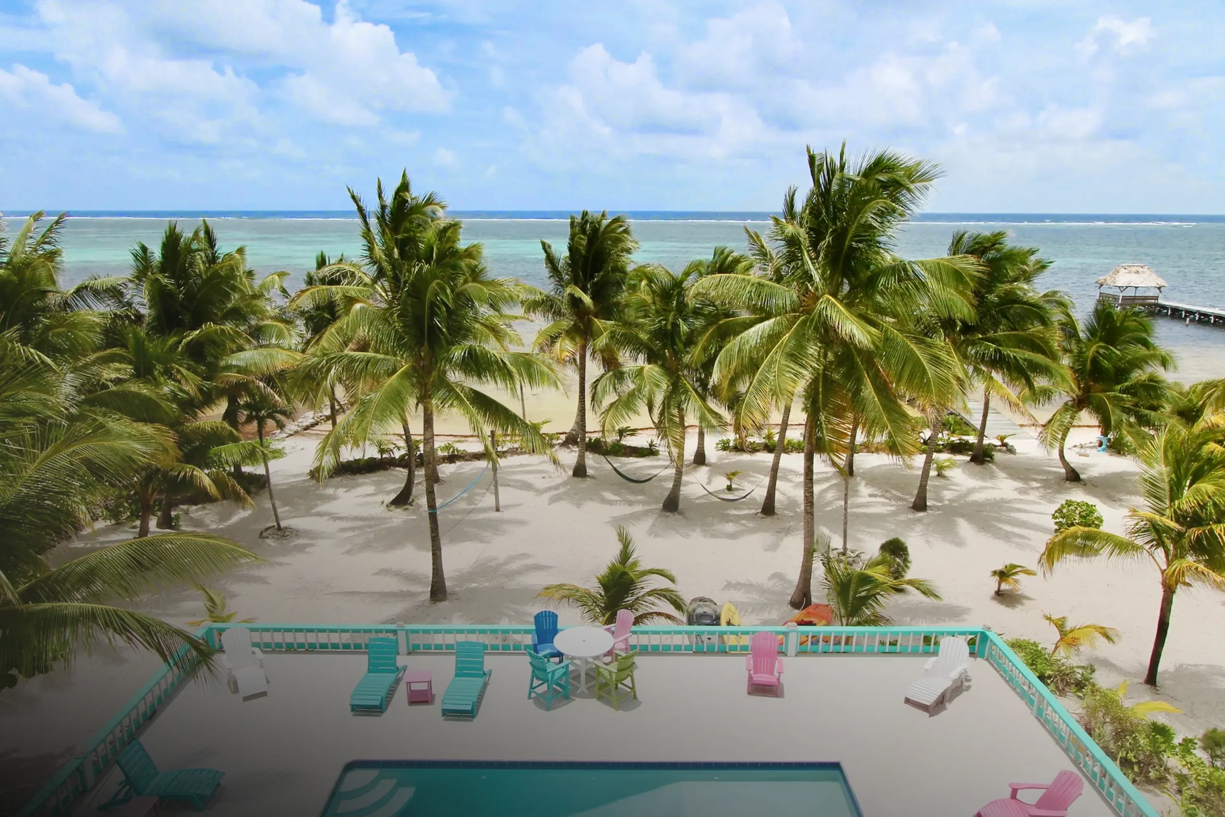 The villa's pool deck and its palms from above, looking past the beach and a thatched pier to the reef