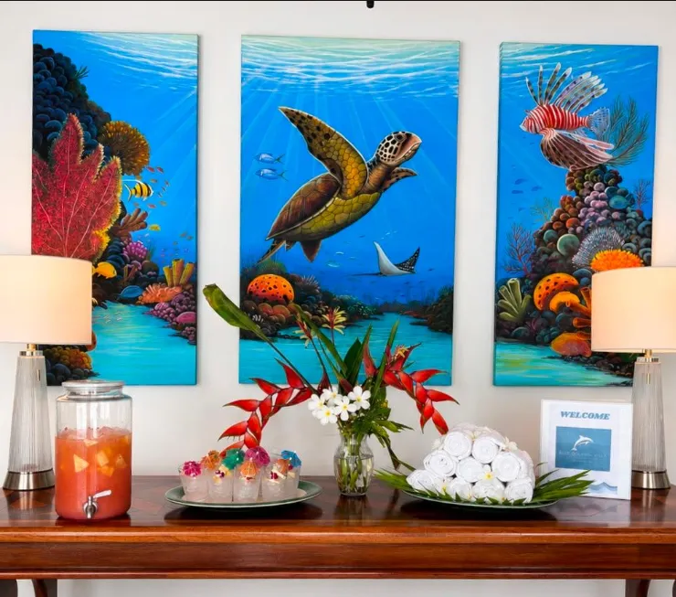 A welcome table under three reef paintings, set with a drinks dispenser, tropical flowers and rolled towels